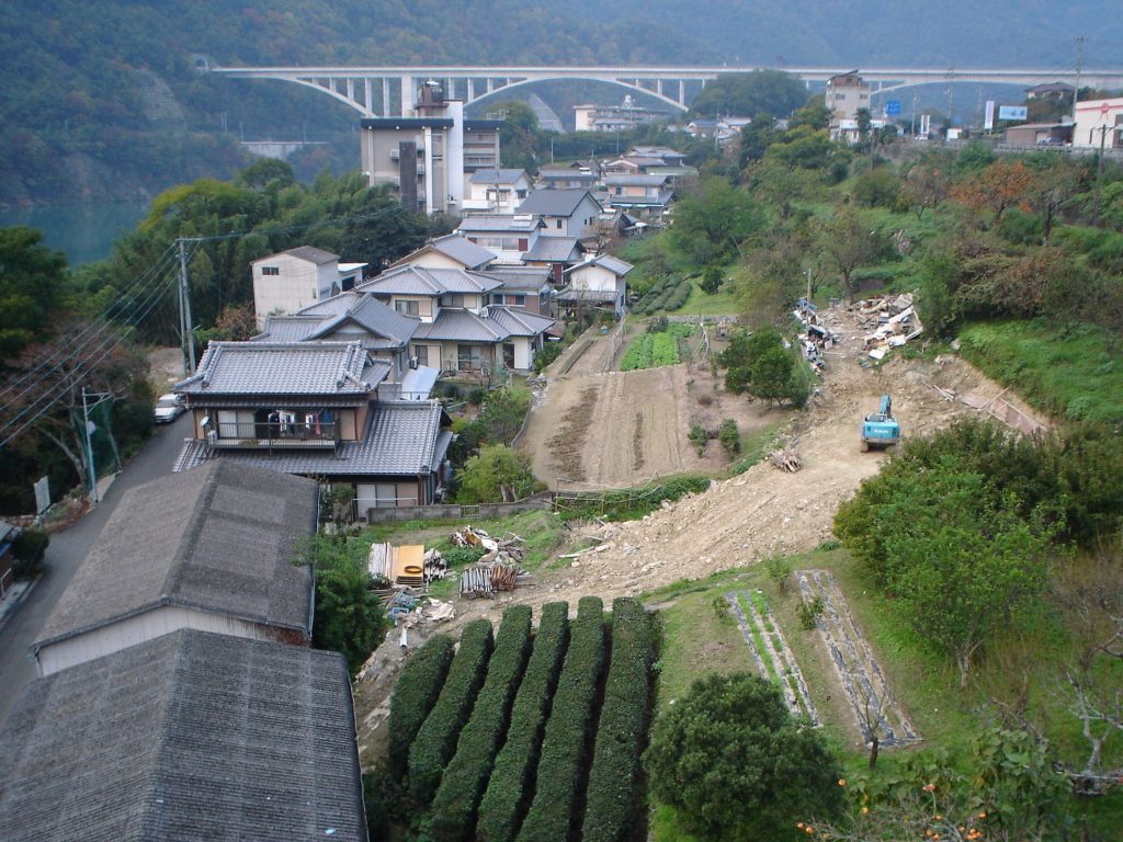 A view of the town of Ikeda, part of Miyoshi City. A series of houses curve from the lower left into the background with a broad arch bridge in the background. There's terraced farmland immediately to the right of the houses. In the background, by the bridge, a river and mountains are visible.